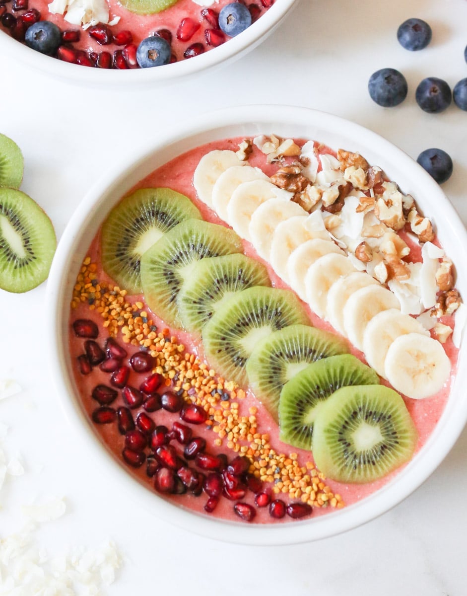 bowl of strawberry kombucha smoothie bowl topped with lines of pomegranate, bee pollen, kiwi slices, banana slices, and shaved coconut.